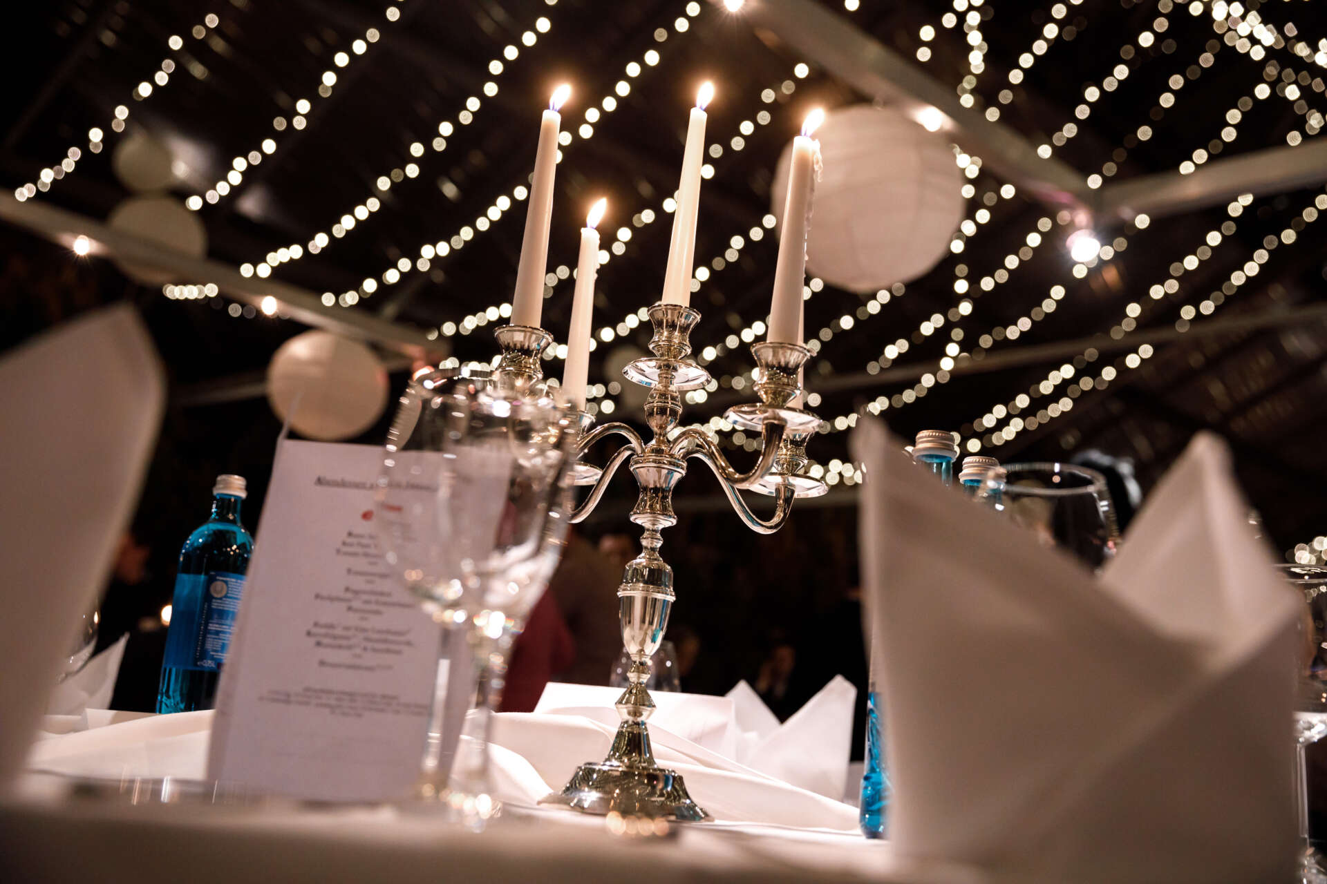 Ein Kerzenstaender steht auf einem weiß gedeckten Tisch und dadrueber haengen Lichterketten