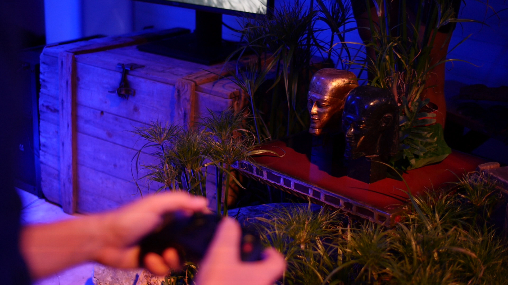 Im Vordergrund wird ein Controller von zwei Haenden gehalten und zwei Buesten und Pflanzen stehen auf einem antiken Hocker neben einem Bildschirm