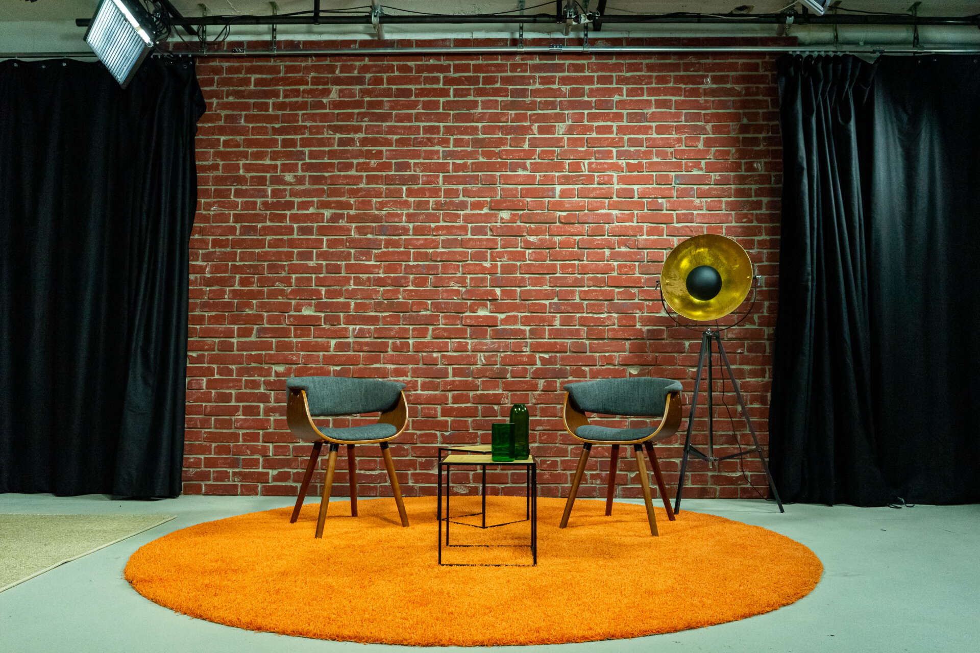 In einem Studio stehen zwei Stuehle, zwei Beistelltische und eine Lampe auf einem orangenen Teppich vor einer Ziegelwand