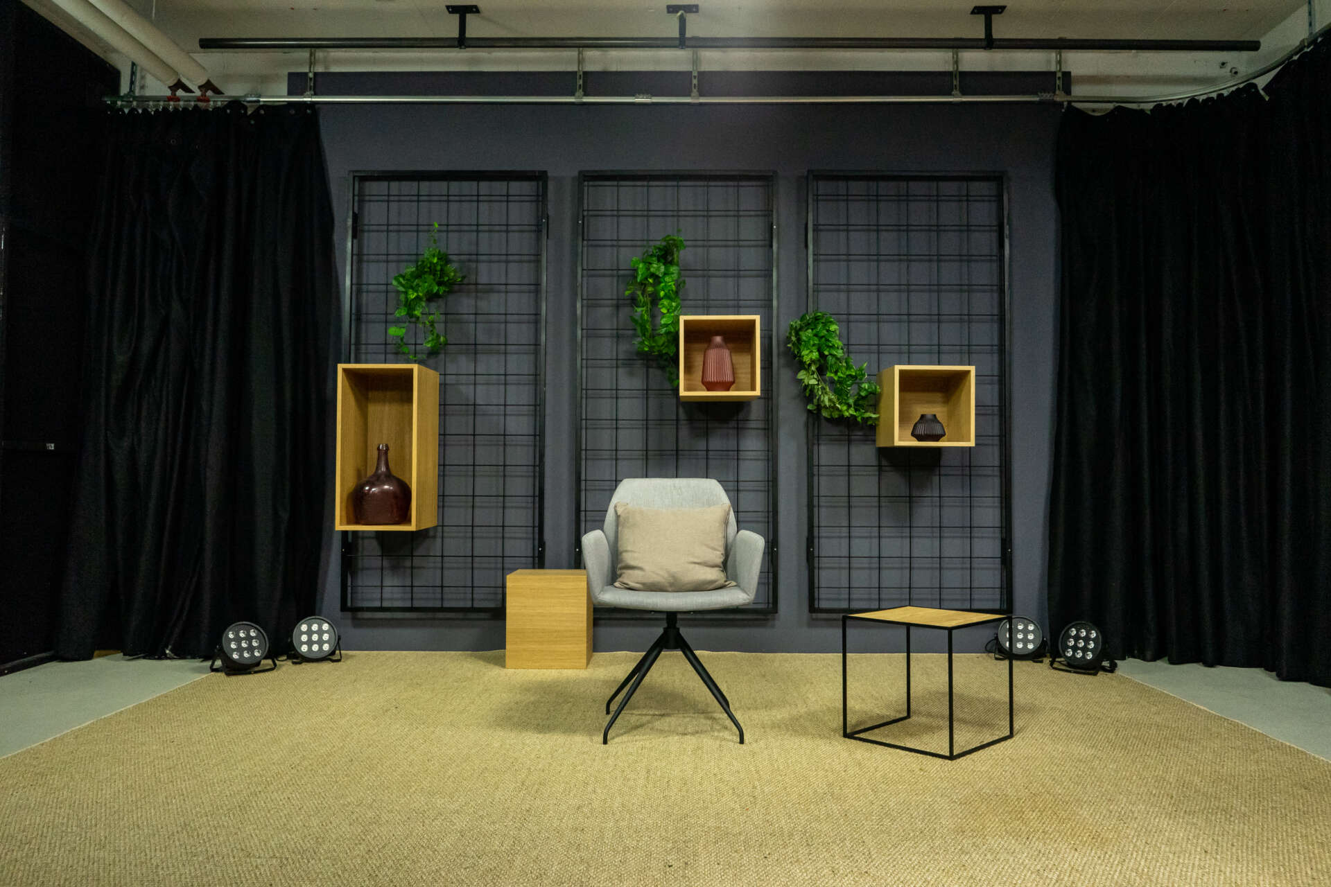 In einem Studio steht ein Stuhl neben einem Beistelltisch auf einem beigen Teppich vor einer Gitterwand, an der drei Kästen und Pflanzen hängen